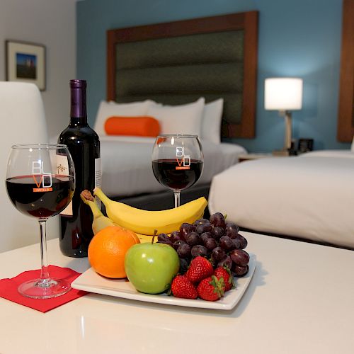 A hotel room with two beds, a table holding a fruit plate, two glasses of red wine, and a bottle of wine on it.