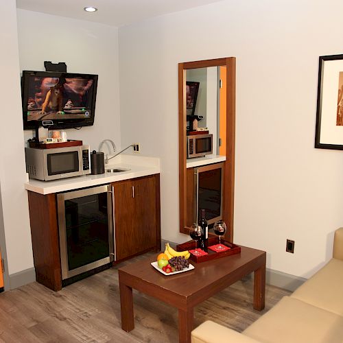 This image shows a cozy room with a kitchenette, a small table with fruits and wine, a sofa, a wall-mounted TV, a mirror, and a framed picture.