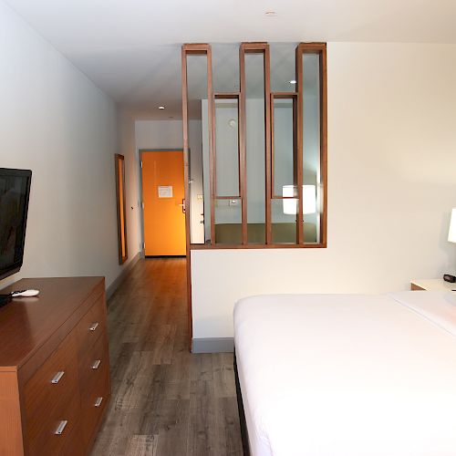 This image depicts a hotel room with a bed, a TV on a dresser, and a bright orange pillow. The room has a partition and a visible door.