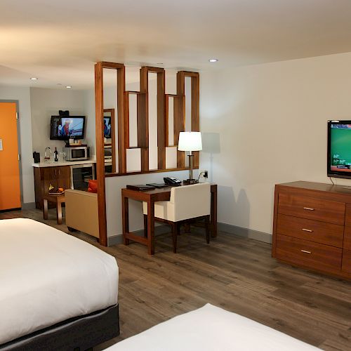 The image shows a hotel room with two beds, a TV, a desk, a chair, and a partial partition. There is a kitchenette area in the background.