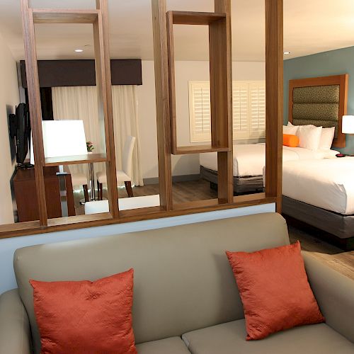 The image shows a hotel room with a beige sofa and orange pillows, a wooden partition, a desk, and two beds with orange accent cushions and blue walls.