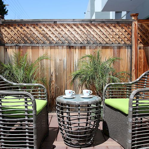 This image shows a small outdoor patio setup with two wicker chairs, a round wicker table, and two coffee cups, all set against a wooden fence.