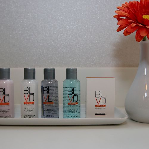 A set of BLVD toiletries including lotion, conditioner, shampoo, and soap next to a vase with an orange flower on a white tray.