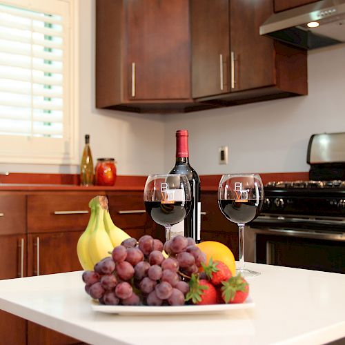 A modern kitchen with a white countertop featuring a fruit platter, wine bottle, and two filled wine glasses, surrounded by wooden cabinets.