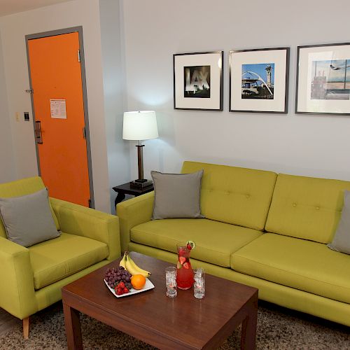 The image shows a living room with a green sofa and armchair, a wooden coffee table with fruits and drinks, and framed pictures on the wall.