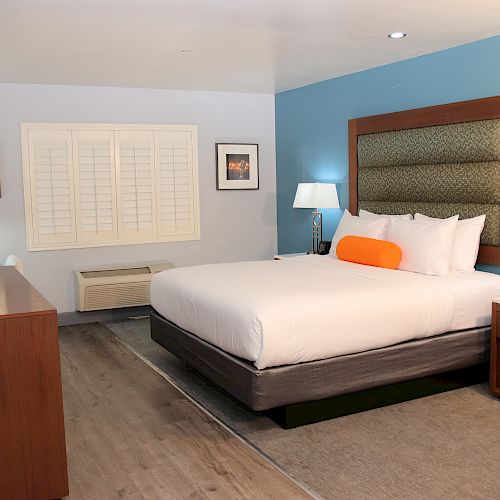 This image shows a hotel room with a large bed, nightstands, lamps, a dresser with a TV, and wall art. The room has blue and white walls.