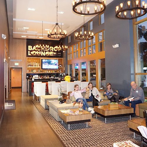 The image shows a modern lounge area with people sitting on sofas, a bar in the background, a TV, and elegant chandeliers above.