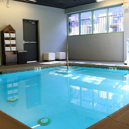 An indoor pool area with a handrail, accessible pool lift, towel rack, seating, and windows allowing natural light to enter the room ending the sentence.