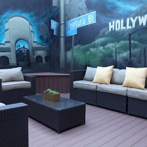 An outdoor patio area with wicker furniture, cushions, and a coffee table; the wall features a Hollywood mural and signs for Ventura Blvd.