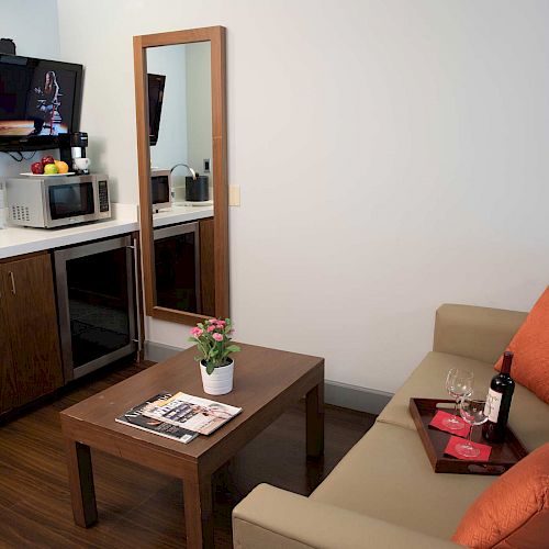 A cozy room with a kitchenette, sofa, table with magazines, wine, and glasses, a TV, and a mirror on the wall. Orange pillows add a pop of color.