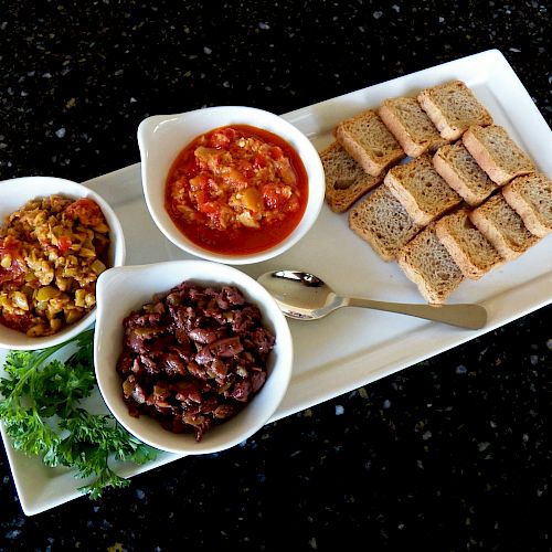 A platter with three bowls: one with a vegetable mix, one with a red stew, and one with black beans, alongside slices of bread and a parsley garnish.