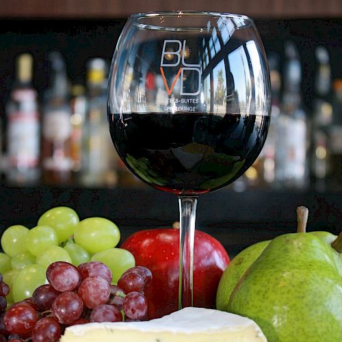 A glass of red wine with cheese, crackers, grapes, an apple, and a pear are displayed on a plate in front of a bar backdrop.