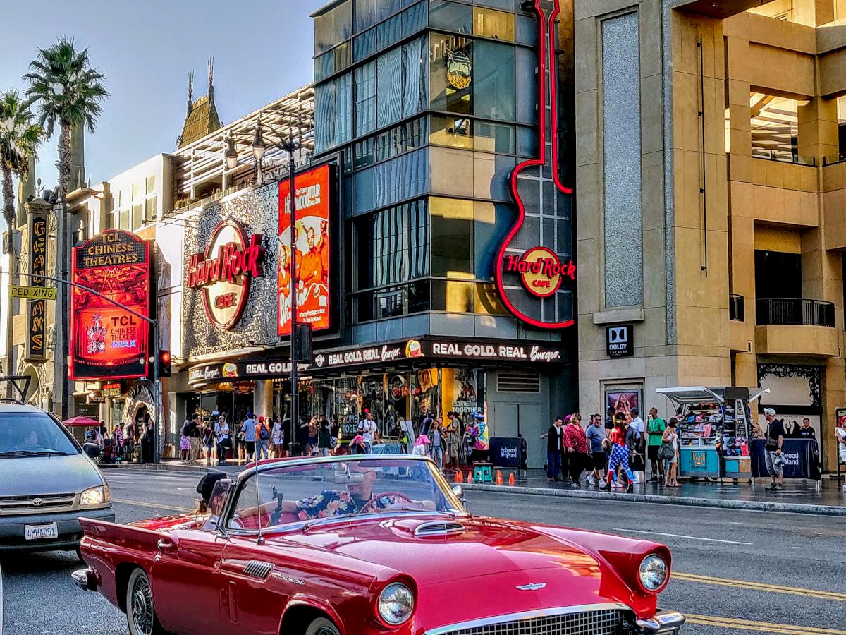 The image shows a red classic convertible car driving in front of buildings with signs and neon lights, people walking nearby, and a sunny sky.