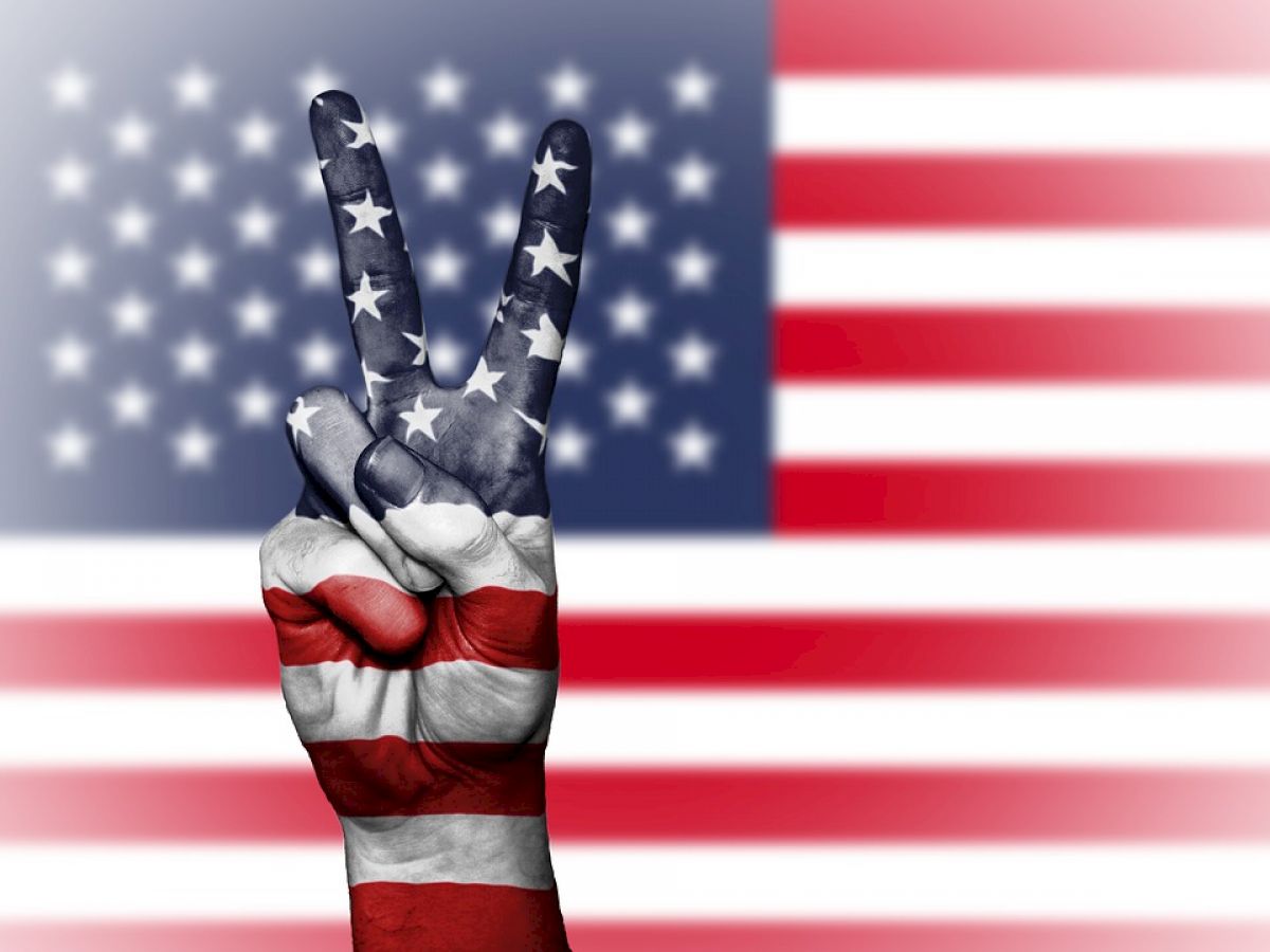 A hand showing a peace sign is painted with the American flag design, set against a background of the American flag.