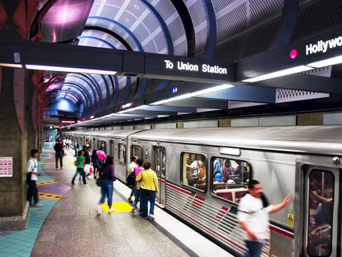 People are at a subway station platform as a train arrives. The signs indicate directions to Union Station and Hollywood.