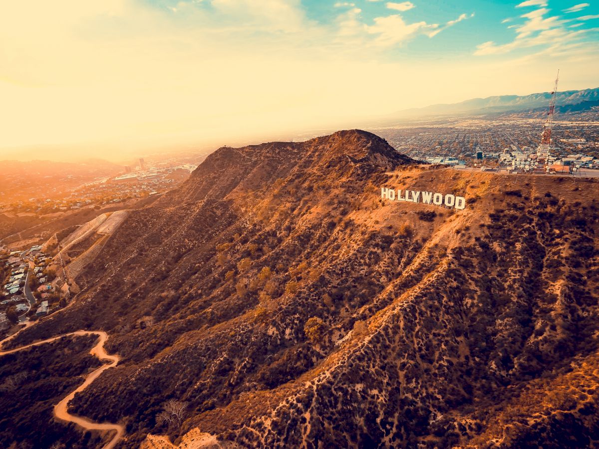 Aerial view of the iconic Hollywood sign on a sunlit hill, overlooking the sprawling city of Los Angeles with mountains and a colorful sky in the background.