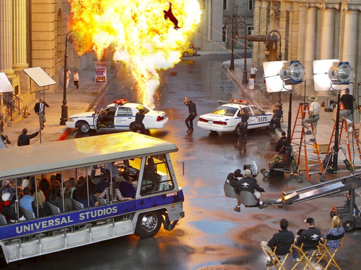 This image shows a movie set with a fiery explosion, police cars, crew members, and a Universal Studios tour tram with spectators.