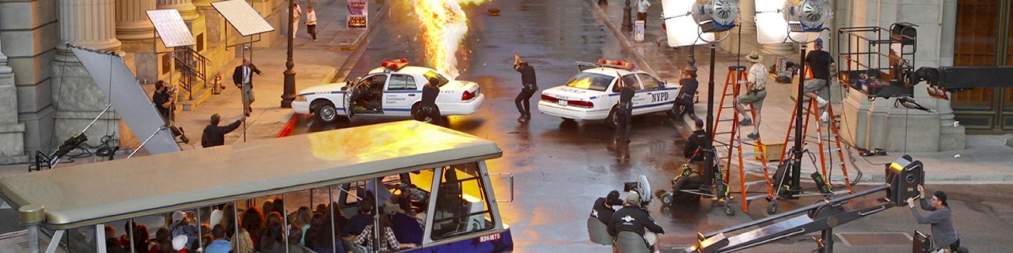 A behind-the-scenes look at a movie set with an explosion, police vehicles, film crew, and a Universal Studios tour tram observing the action.