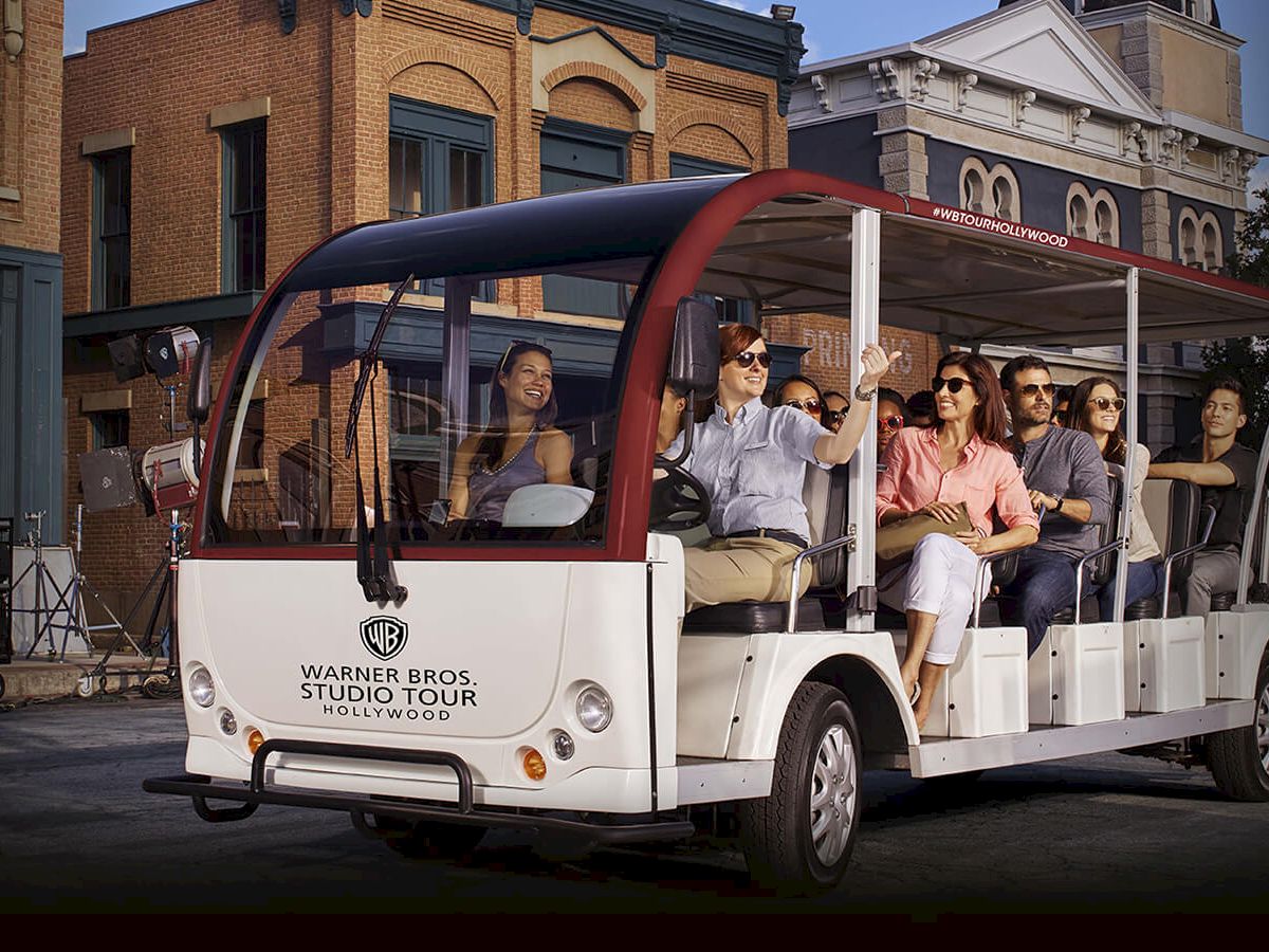 A group of people enjoying a Warner Bros. studio tour in an open-air vehicle, passing by buildings resembling a movie set city backdrop.