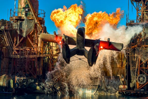 A dramatic scene shows a seaplane taking off with explosions and water splashing around an industrial structure in the background.