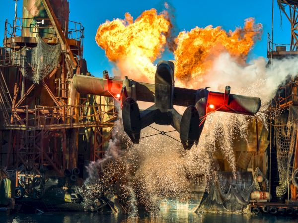 A dramatic scene shows a seaplane taking off with explosions and water splashing around an industrial structure in the background.