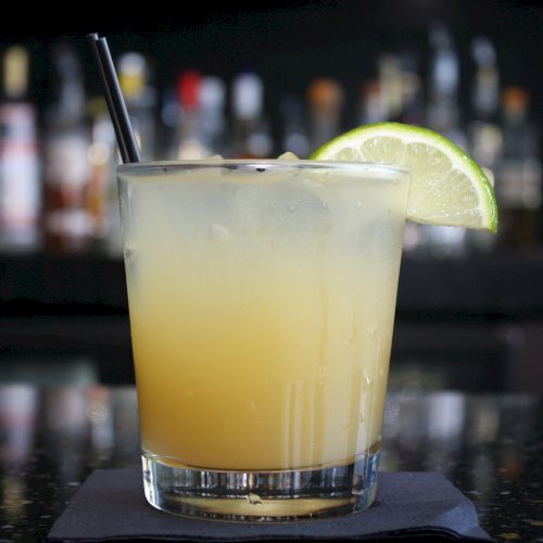 A glass of a yellow cocktail with a black straw and a lime slice garnish, placed on a black napkin with a blurred bar background.