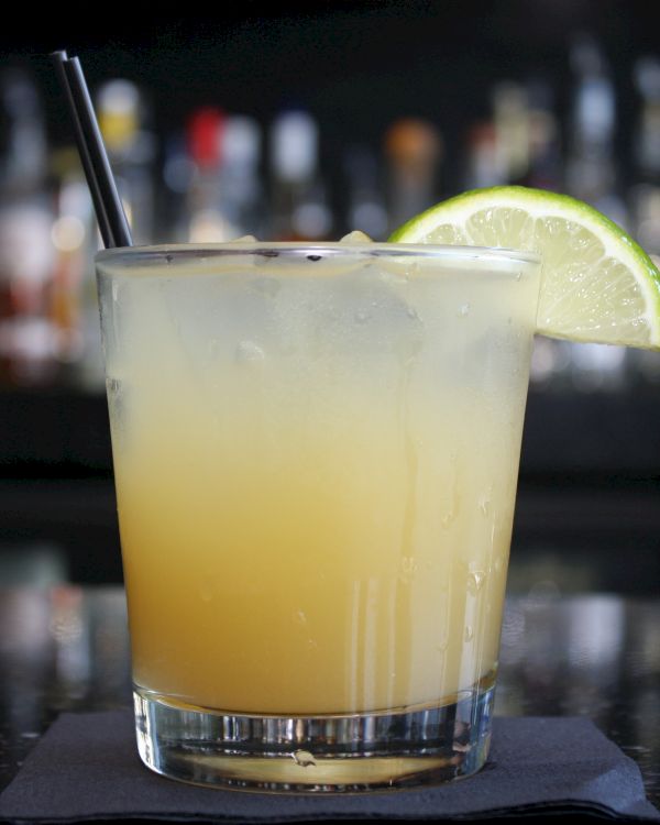 A glass of a yellow cocktail with a black straw and a lime slice garnish, placed on a black napkin with a blurred bar background.