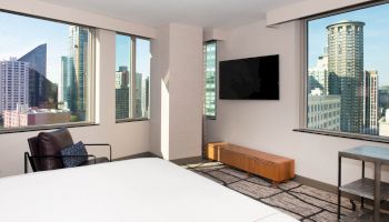 A modern hotel room with large windows showcasing city views, a bed, a chair, a television, and a small table and bench against the walls.