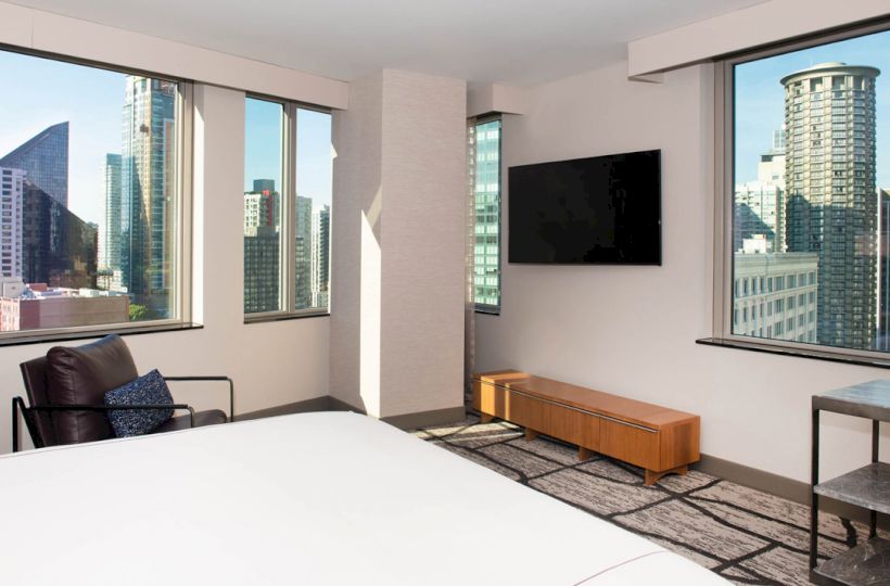 A modern hotel room with large windows showcasing city views, a bed, a chair, a television, and a small table and bench against the walls.