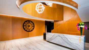 A modern lobby with a marble reception desk, large wooden spiral staircase, round wall clock, and decorative lighting fixtures, plus flowers.