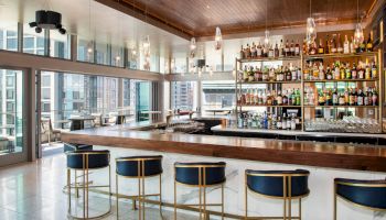 A modern bar with a variety of bottles displayed on shelves behind it, with seating and large windows offering a view of the city outside.