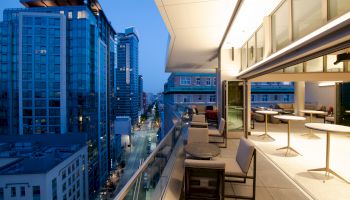 A modern rooftop terrace with seating, overlooking a cityscape with tall buildings as the evening approaches and city lights start to illuminate.