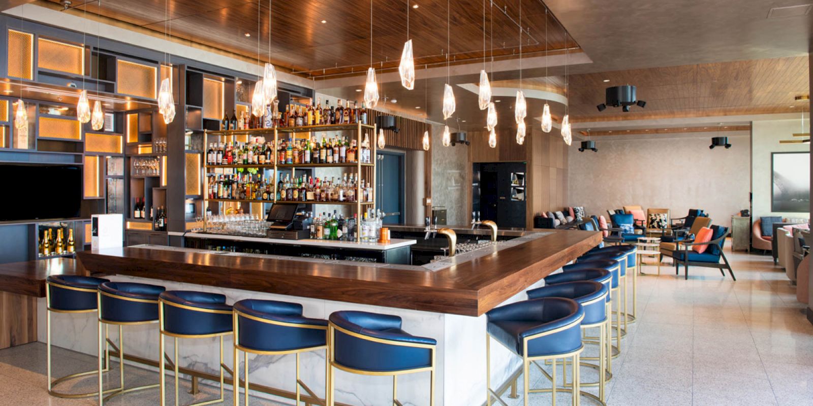 A modern bar with counter seating, hanging pendant lights, and a fully stocked shelf, set in a stylish, spacious lounge area.