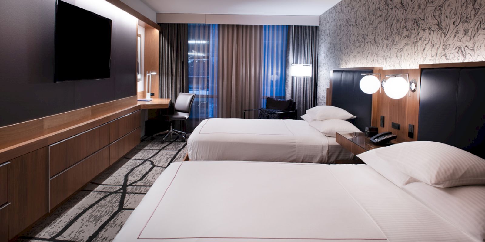 The image shows a modern hotel room with two neatly made beds, a wall-mounted TV, a desk with a chair, and a window with curtains at the back.