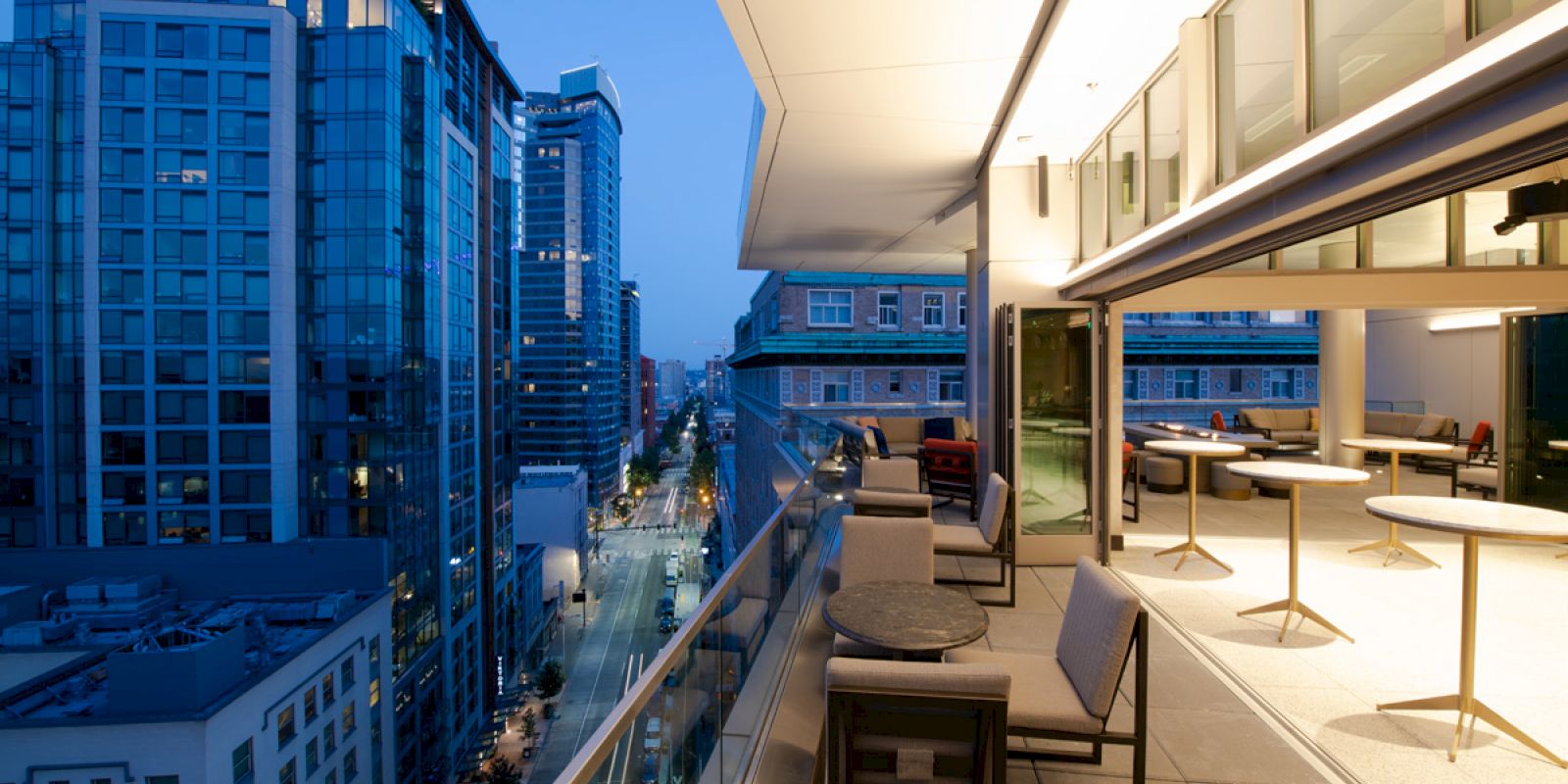 A modern urban balcony with seating area, overlooking a city street and high-rise buildings at dusk. Indoor and outdoor spaces blend seamlessly.