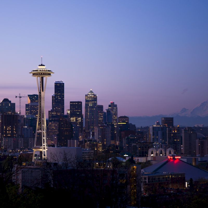 A cityscape view of Seattle at twilight, featuring the Space Needle and distant Mount Rainier, surrounded by numerous buildings.