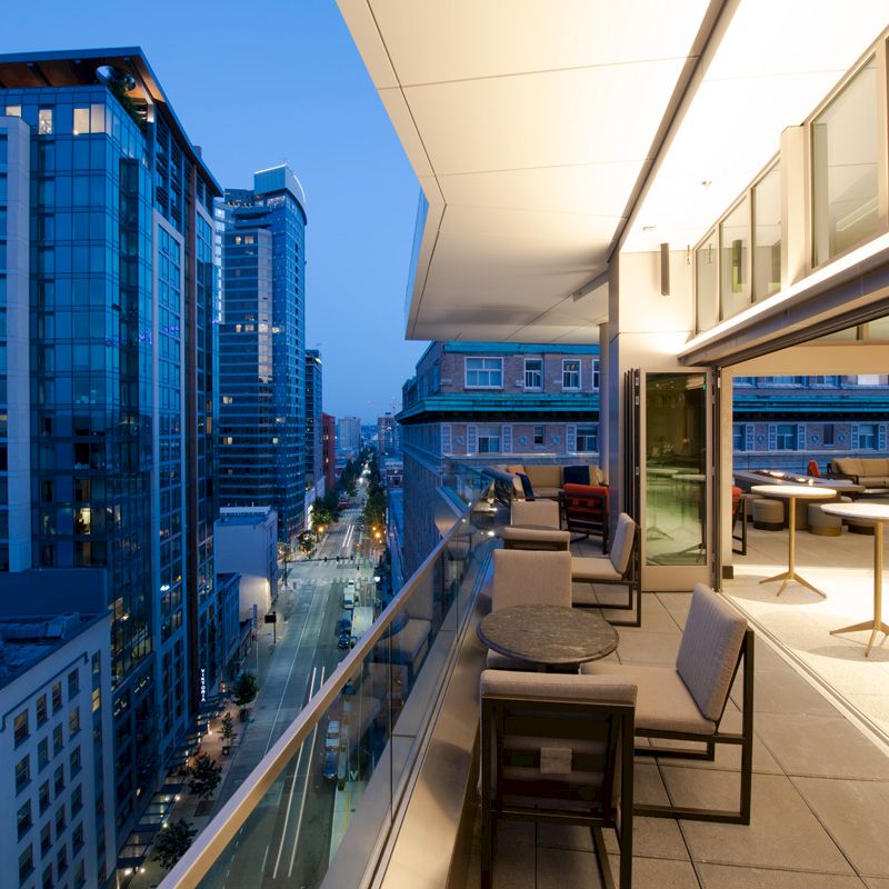 This image shows a modern cityscape with tall buildings at dusk. In the foreground is a stylish patio with seating and tables, attached to a building.
