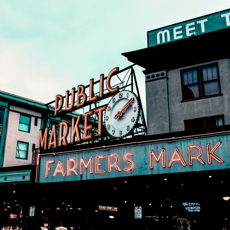 The image shows a neon-lit sign for a public farmers market, with a clock and adjacent buildings, creating an urban market atmosphere.
