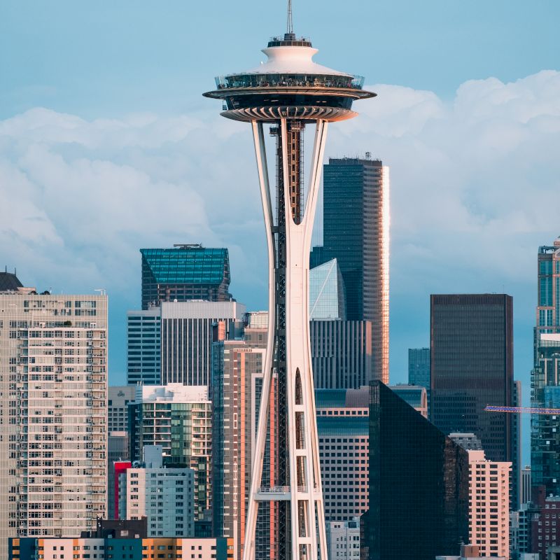 The image shows the Space Needle in Seattle, Washington, with modern skyscrapers and a cloudy sky in the background, an iconic landmark of the city.