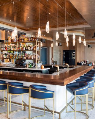 The image shows a modern bar with a wooden counter, blue bar stools, and hanging lights. The well-stocked bar has various bottles, glasses, and seating.