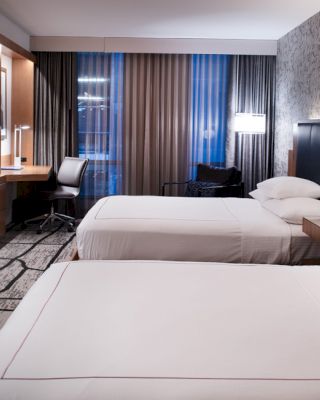 The image shows a modern hotel room with two neatly made beds, a flat-screen TV, a desk with a chair, and floor-to-ceiling windows with curtains.