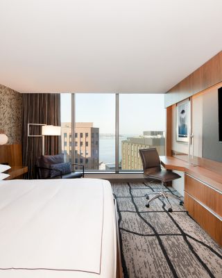 A modern hotel room featuring a large bed, a wall-mounted TV, a desk with a chair, and a city view from a large window at the far end.