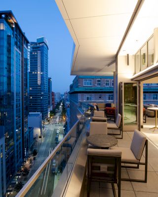 An outdoor terrace with tables and chairs overlooks a city street lined with high-rise buildings in the evening.