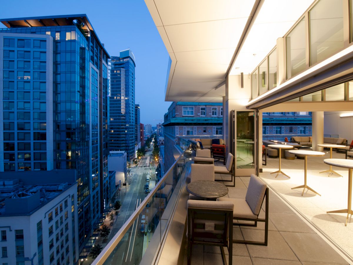 A modern cityscape during evening with tall buildings, accompanied by a well-furnished balcony featuring tables and chairs overlooking the street below.