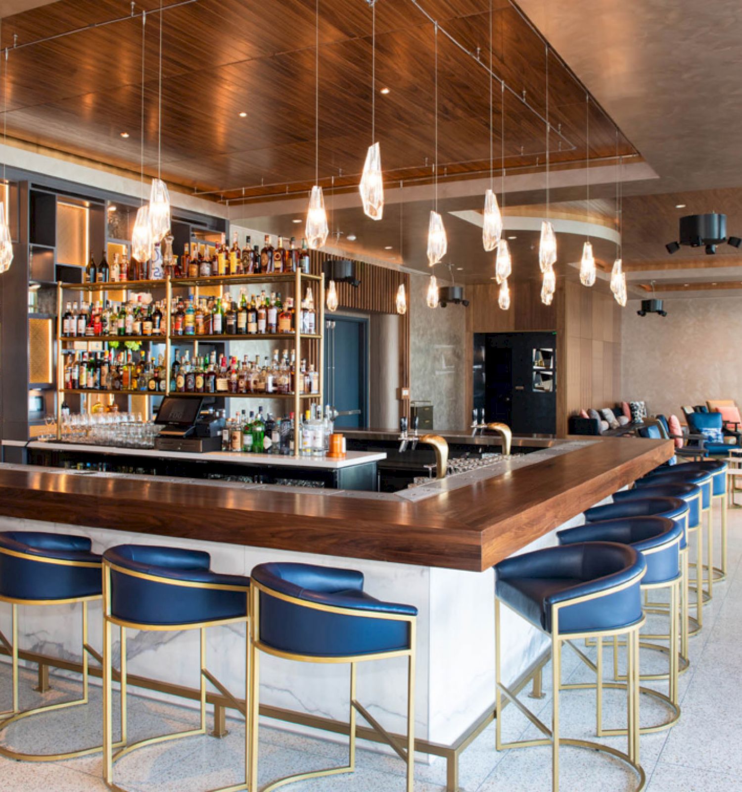 A modern bar with a central island, blue barstools, a variety of liquor bottles, pendant lights, and contemporary seating in the background.