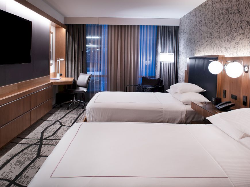 The image shows a modern hotel room with two double beds, a flat-screen TV, a desk with a chair, lamps, and large windows with curtains.