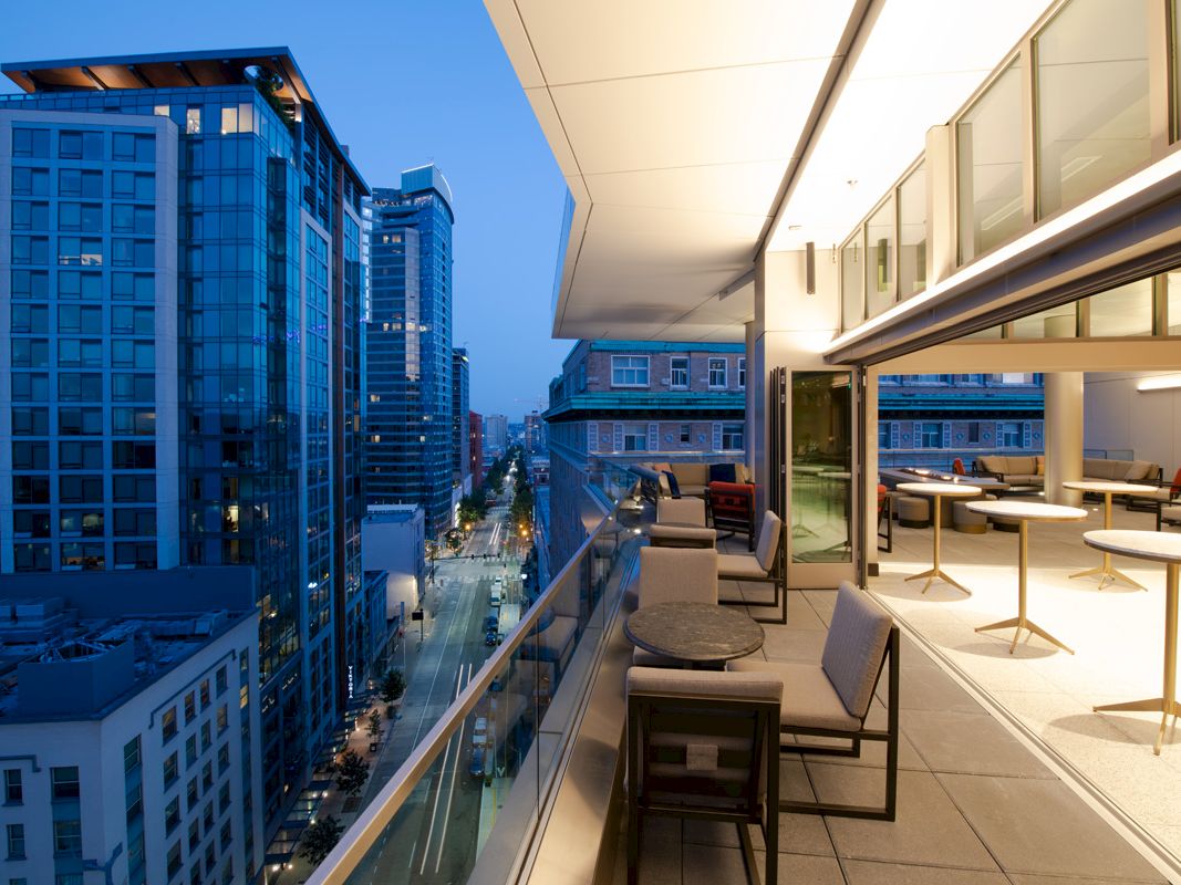 A modern urban balcony at dusk with tables and seating, overlooking a city street lined with tall, illuminated buildings.