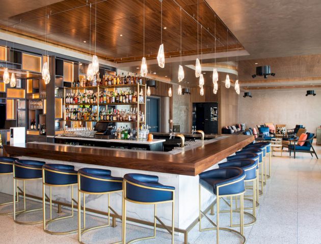 The image shows a modern bar with blue and gold stools, a wooden bar counter, hanging lights, and a fully stocked bar. The area looks spacious.