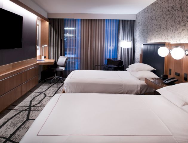A modern hotel room with two beds, a flat-screen TV, a desk with a chair, and stylish decor, including a patterned wall and carpet.
