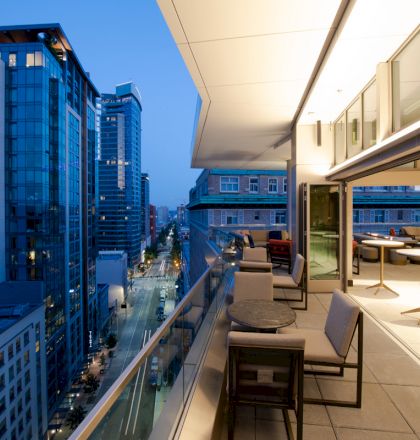 It's an urban balcony at dusk, with a view of high-rise buildings and a street below; it features chairs, tables, and a glass railing.